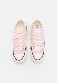 Růžové plátěné tenisky s bílými tkaničkami, gumovou špičkou a černým akcentním pruhem. Na jazyku mají značku "Converse All-Star".