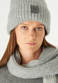Bonnet en tricot gris avec une texture côtelée et un patch logo en tissu gris, associé à une écharpe douce grise présentant une texture similaire.