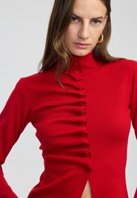 Czerwony długi rękaw top z wysokim kołnierzem, zapinany z przodu na guziki z falbankami oraz z bocznym rozcięciem. Materiał ma delikatną żebrowaną strukturę.