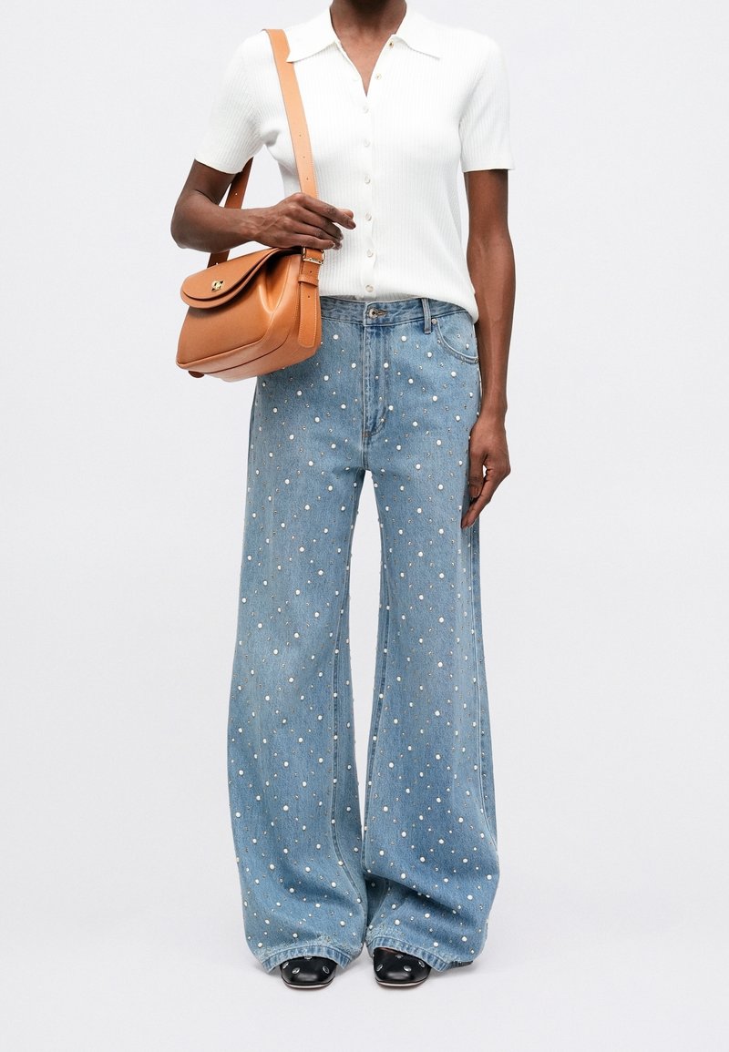 Personne portant un haut blanc à manches courtes boutonné, un jean large bleu clair à pois blancs, des chaussures noires, et portant un sac à épaule marron.