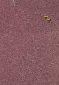 Tessuto color marrone scuro con piccolo alce marrone ricamato con corna gialle vicino all'angolo in alto a destra e parziale cerniera argentata al centro in alto.