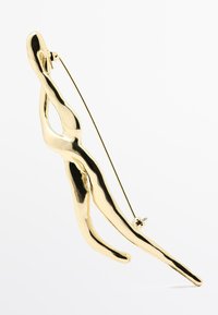 Spilla in tono oro con un design a figura umana astratta, presentante una texture levigata e liscia e una spilla sul retro per il fissaggio.