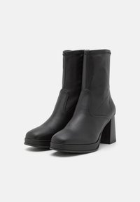 Une paire de bottes en cuir noir mi-mollet avec des talons blocs et des pointes arrondies, placées côte à côte sur un fond blanc uni.