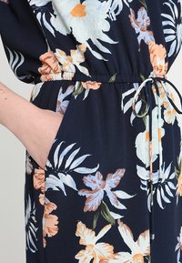 Květinový overal s námořnickým pozadím, lehkým materiálem, elastickým pasem s šňůrkou a bočními kapsami zdobenými květinami a listy.