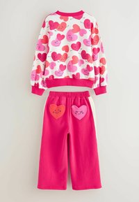 Set format din bluză și pantaloni, realizat din țesătură moale. Bluza albă cu modele de inimioare roz/roșii; pantalonii roz cu două buzunare în formă de inimă, unul roșu și unul roz.
