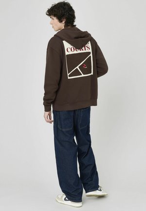 Haut à capuche marron avec un grand motif crème et brun foncé au dos portant le mot "COURTS". Porté avec un jean bleu et des baskets blanches.