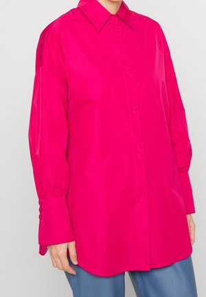 Kobieta w jasnoróżowej, długorękawowej koszuli zapinanej na guziki z szerokim kołnierzem oraz niebieskich spodniach, stojąca na tle jednolitego tła.