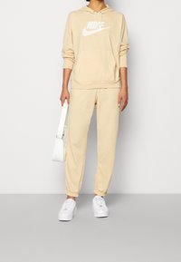 Beige mikina Nike s bílým logem, v kombinaci s odpovídajícími béžovými tepláky. Obsahuje volnější střih, kapuci se stahovací šňůrkou a elastické manžety. Bílé tenisky.