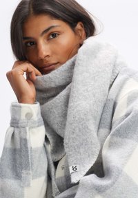 Écharpe en laine à carreaux gris et blancs avec une finition douce et texturée ; présente des bords roulés et une petite étiquette de marque sur le côté.