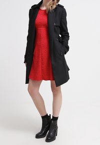 Svart trench coat med dubbelknapprad, bärs över en texturerad röd klänning. Svarta ankelskor med sidodragkedja och kraftig klack.