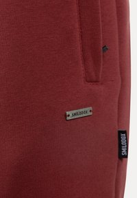 La tela de mezcla de algodón roja presenta una etiqueta de metal "SMILODOX". Los detalles notables incluyen un bolsillo lateral y una etiqueta tejida en el dobladillo.