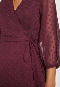 Abito avvolgente color bordeaux con texture a pois, maniche trasparenti e fiocco laterale. Presenta una scollatura a V profonda e una forma A-line.