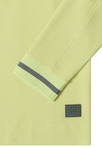 Chemise à manches longues vert clair avec une bande grise sur le poignet et une petite étiquette logo carrée noire à l'ourlet.