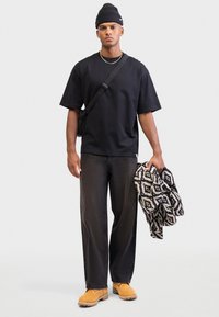 Crna kratka majica s kratkim rukavima, tamne traperice, žute čizme, drži uzorčani crno-bijeli pokrivač, nosi pletenu kapu i torbu preko tijela.