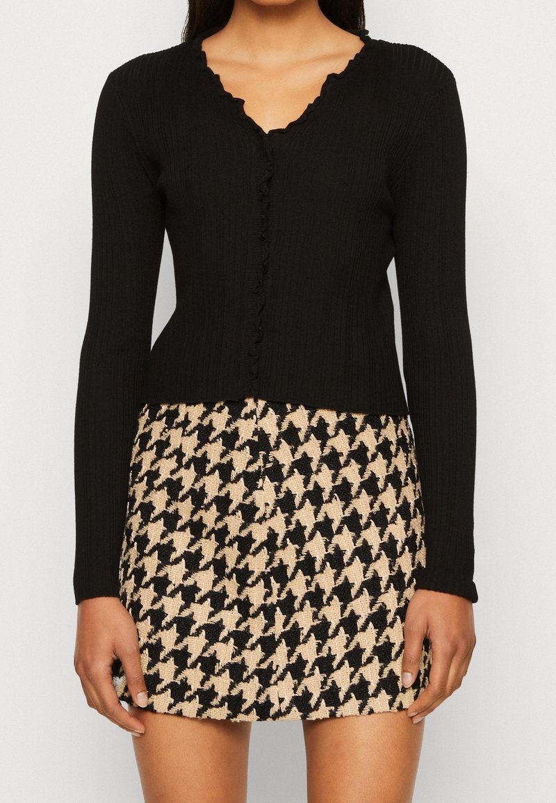 Femme portant un cardigan noir à manches longues en tissu côtelé avec des bords volantés et une jupe à motif pied-de-poule beige et noir.