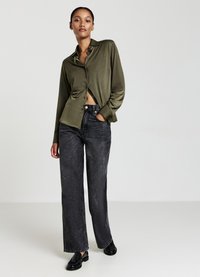 Camicia a bottoni in satin verde oliva, infilata davanti, abbinata a jeans neri a vita alta e gamba larga e mocassini neri. Tessuto morbido, vestibilità rilassata.