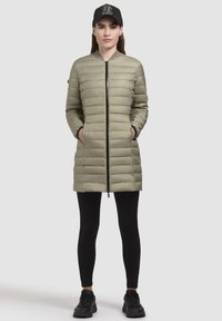 Fodrad olivgrön jacka med frontdragkedja, ribbad krage, två sidofickor, bärs över svarta leggings och sneakers, svart keps med logotyp.