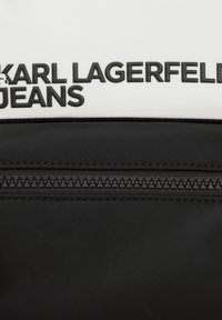 Zaino bianco e nero realizzato in un materiale nylon liscio, con testo in rilievo "KARL LAGERFELD JEANS" e una cerniera nera sulla tasca frontale.