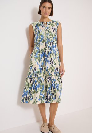 Kvinde stående iført en ærmeløs midi-kjole med blåt, grønt og cremefarvet abstrakt blomsterprint og beige slip-on sko.