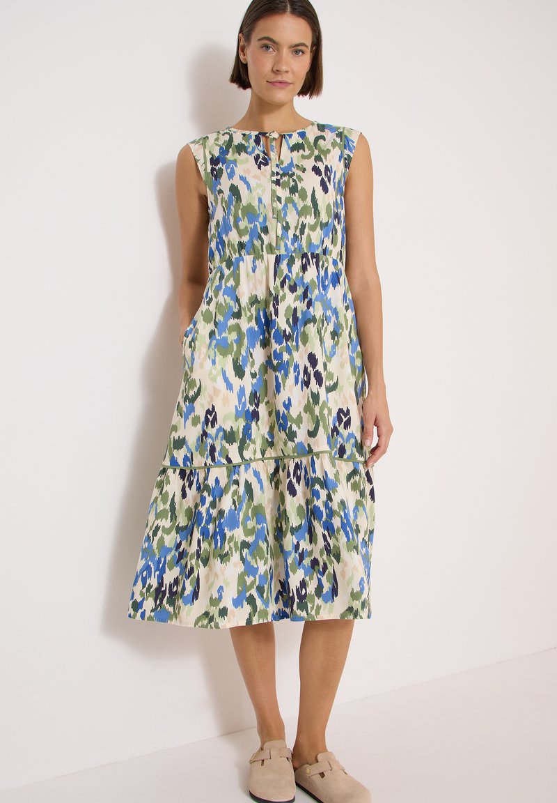 Femme debout portant une robe midi sans manches avec un imprimé floral abstrait bleu, vert et crème et des chaussures beige sans lacets.