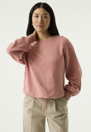 Vrouw met lang donker haar draagt een losse roze trui en beige broek, staat met één hand aan haar nek, naar voren gericht.