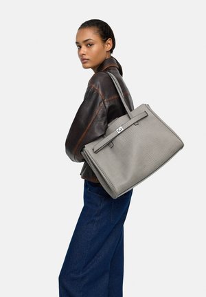 Kobieta w ciemnobrązowej skórzanej kurtce i niebieskich spodniach niesie przez ramię dużą, szarą, teksturowaną torebkę na białym tle.