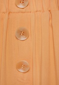 Meruňková látková košile s třemi velkými, průhlednými knoflíky. Látka má jemnou texturu a subtilní diagonální žebrování.