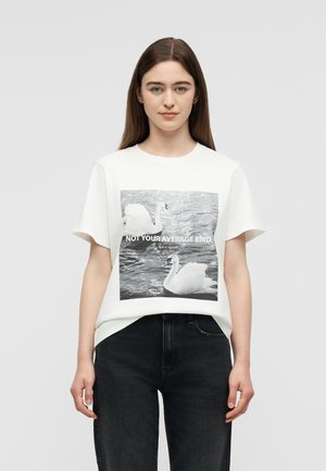 Młoda kobieta z długimi brązowymi włosami, ubrana w białą koszulkę, pokazująca dwa łabędzie na wodzie oraz napis "Nie twój przeciętny ptak."