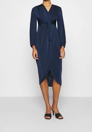 Femme portant une robe midi portefeuille bleu marine à manches longues avec ceinture à la taille, associée à des sandales noires à talons ouvertes.