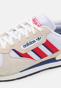 Tenisky Adidas s béžovým semišovým a bílým síťovaným svrškem, červenými a modrými pruhy a bílým zavazováním na tkaničky s logem.
