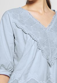 Světle modrý tunikový top s dekorativním výstřihem z broderie anglaise, krátkými nabíranými rukávy a uvolněným střihem s vyšívanými detaily.