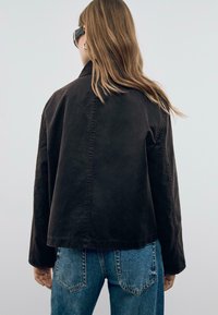 Veste marron foncé à coupe décontractée, avec un col Mandarin et des coutures visibles, associée à un jean bleu taille haute.