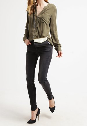 Vrouw draagt een olijfgroene blouse met knopen, zwarte skinny jeans, zwarte hoge hakken en een kleine zwarte en gouden schoudertas.