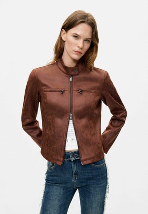 Jeune femme portant une veste en cuir marron zippée et un jean bleu, debout avec les mains dans le dos, expression neutre, fond blanc.
