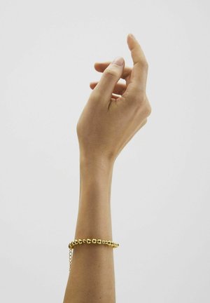 Raised hand with wrist adorned by a gold beaded bracelet against a plain light background.
