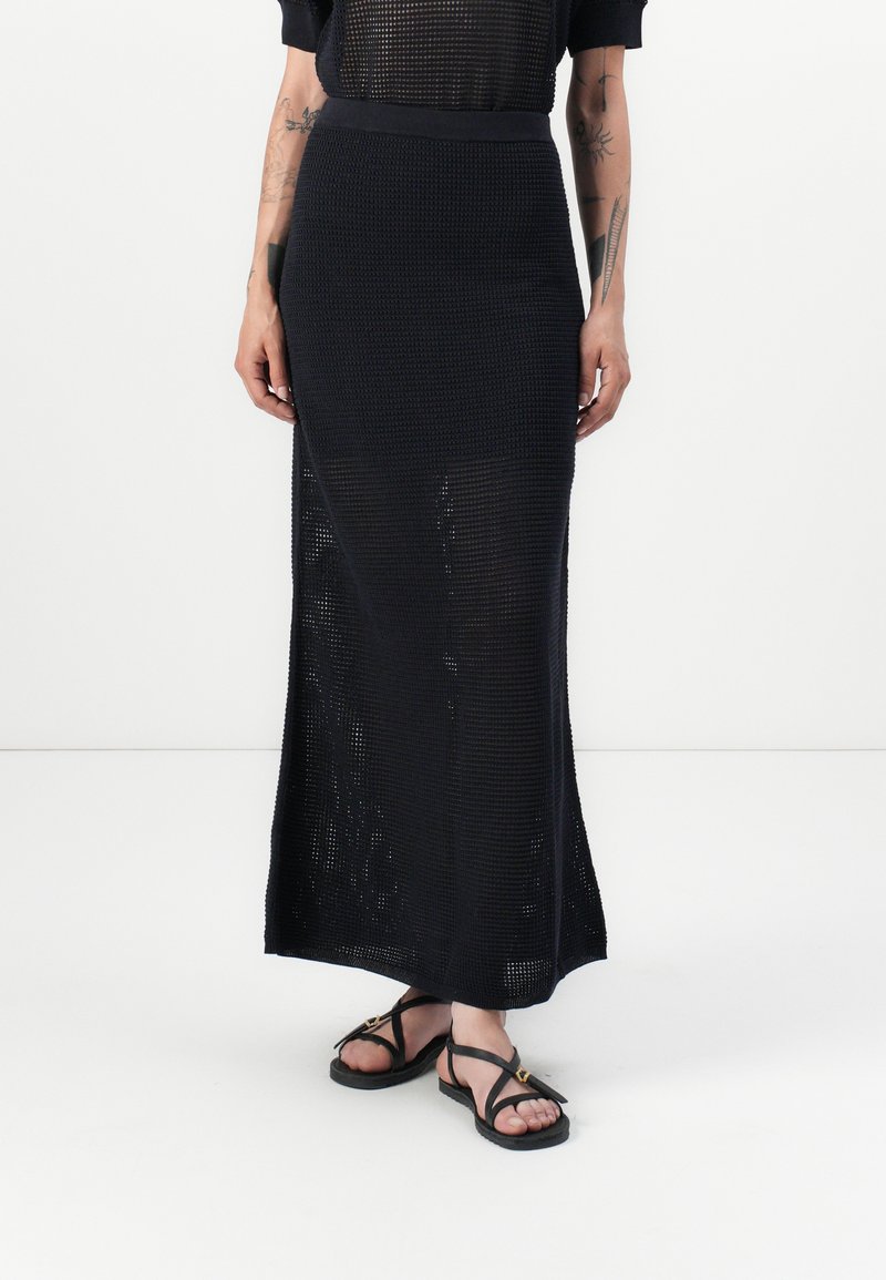 Zwarte gebreide maxi-rok met een geribbelde textuur, hoge tailleband en zijknielen. Gepaard met platte zwarte sandalen met gouden accenten.