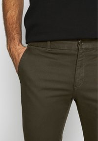 Spodnie w kolorze oliwkowej zieleni o teksturowanym splocie, z przodu kieszeń, zapięcie na guzik oraz wąski krój, na neutralnym tle.