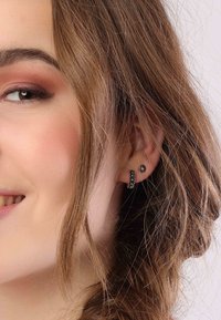 Zwarte studs en hoepeloorbellen met kleine studaccenten. De hoepel heeft een geprofileerd ontwerp. Het haar is golvend en valt rond het gezicht.