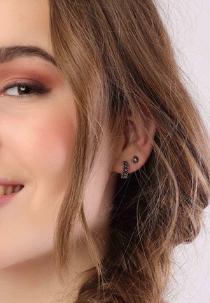 Zwarte studs en hoepeloorbellen met kleine studaccenten. De hoepel heeft een geprofileerd ontwerp. Het haar is golvend en valt rond het gezicht.