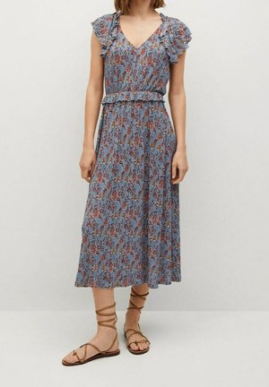 Vrouw draagt een blauwe midi-jurk met bloemen en ruches aan de mouwen en taille, gecombineerd met bruine veterplatte sandalen, staand tegen een effen achtergrond.