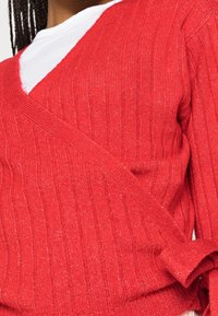 Suéter rojo de punto con textura acanalada, que presenta un escote en V y un diseño cruzado. Fabricado con hilo suave y con sutiles hilos metálicos.