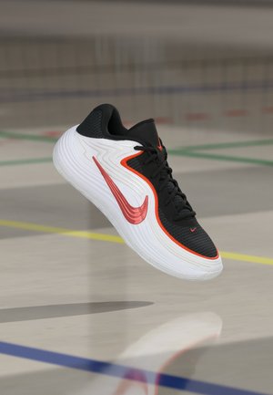 Fekete-fehér sportcipő piros kiemelésekkel és pipa logóval, egy konditerem padlója felett lebeg, amelyen pályajelölések láthatók.