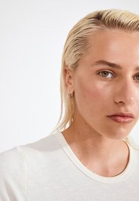 Detailný záber na osobu s svetlobéžovým rebrovaným tričkom. Vlasy sú slicked back a vidieť malé strieborné kruhové náušnice. Pokožka vyzerá hladko.