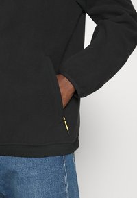 Černý fleecový pulovr se zipovou kapsou, elastickými manžetami a texturovaným povrchem. V kombinaci s modrými džíny.