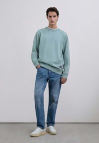 Sudadera verde clara con cuello y puños de canalé, combinada con jeans de mezclilla azul claro y zapatillas blancas con suelas amarillas.