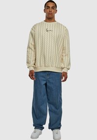 Sudadera color crema con patrones negros ondulados, puños acanalados y cuello redondo. Combinada con jeans azules holgados y zapatillas blancas.