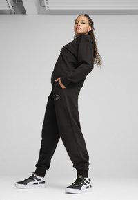 Juodas sportinis megztinis ir kelnių komplektas, pasižymintis laisvu siluetu, šoninėmis kišenėmis ir logotipu. Batai juodi ir balti, su masyviu dizainu.