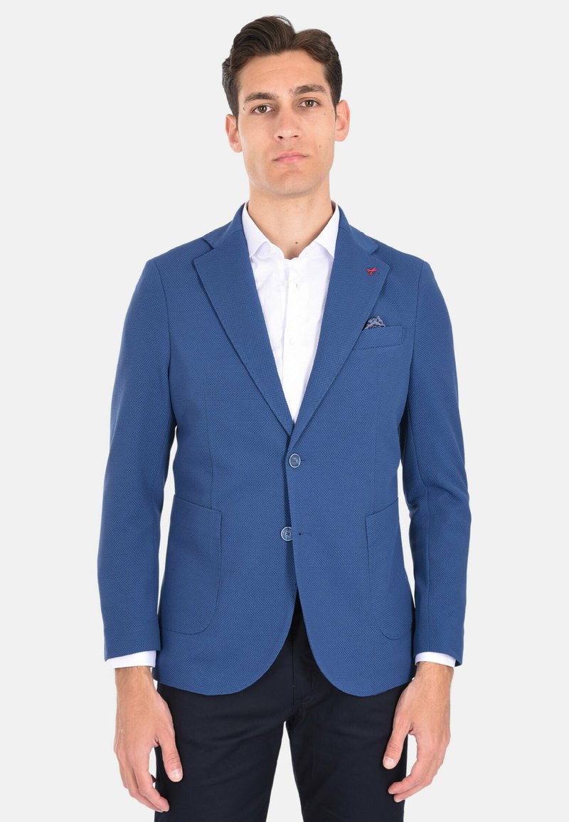 Blazer blu con texture, dotato di due tasche frontali, due bottoni e un piccolo emblema rosso. Indossato sopra una camicia bianca, con un fazzoletto da taschino.