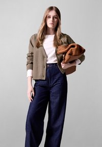 Olivgrüner, gestrickter Cardigan mit gerippten Bündchen, kombiniert mit einem weißen T-Shirt und hoch taillierten, dunkelblauen, weit geschnittenen Jeans. Sie hält eine braune Tasche.