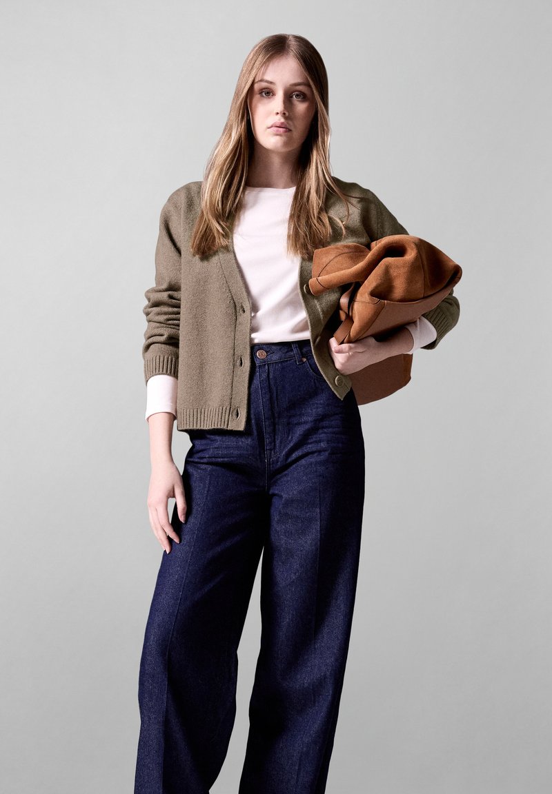 Olivgrüner, gestrickter Cardigan mit gerippten Bündchen, kombiniert mit einem weißen T-Shirt und hoch taillierten, dunkelblauen, weit geschnittenen Jeans. Sie hält eine braune Tasche.